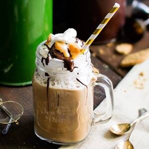 May include: A glass mason jar filled with a brown iced coffee drink topped with whipped cream, chocolate sauce, and peanut butter cups. A gold and white striped straw is in the drink.