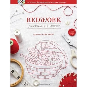 Pode incluir: Um livro intitulado "REDWORK from TheWORKBASKET" com materiais de bordado. A capa apresenta uma ilustração de uma cesta cheia de linha, tesoura e agulhas. Outros itens incluem uma fita métrica, botões e uma almofada de alfinetes vermelha.