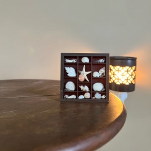 May include: A dark brown, square shadow box filled with various seashells and a starfish. The box is divided into multiple compartments, each holding a different type of shell. A decorative plug-in wax warmer is in the background.