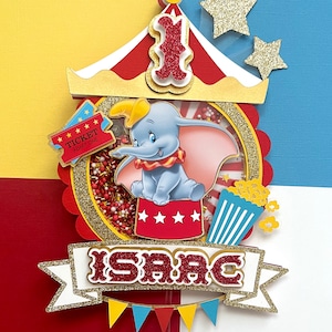 May include: A red, white, and gold circus-themed cake topper with a Dumbo the Elephant character, a red and white striped tent, a popcorn bucket, and a banner that says "ISAAAC".