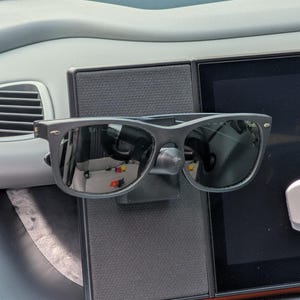 Pode incluir: Óculos de sol pretos montados no painel de um carro. Os óculos de sol são presos por um suporte preto. O painel é cinza e possui um visor digital.