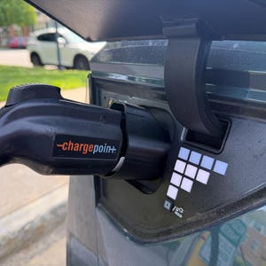 Può includere: Una maniglia di ricarica per veicoli elettrici nera con il logo ChargePoint inserita in una porta di ricarica per auto. L'area della porta presenta un alloggiamento in plastica nera con una griglia bianca di luci quadrate e un'icona HOLD.