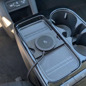 Pode incluir: Um carregador sem fio Qi preto instalado na consola central de um carro. A almofada é redonda com o logotipo Qi e um cabo conectado. A consola tem uma superfície texturizada e um porta-copos.
