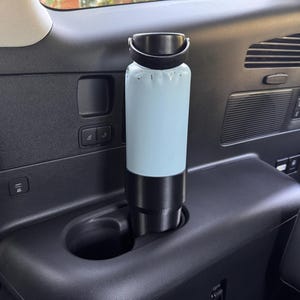May include: A light blue water bottle with a black base and handle, placed in a car's cup holder. The bottle has a matte finish and a black lid. The cup holder is black, and the interior of the car is visible in the background.
