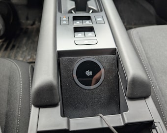 May include: A black wireless charging pad with a circular Qi2 logo and 15W text integrated into a vehicle center console. The charging unit is positioned between two gray textured armrests, featuring a sleek matte finish and a thin power cable.