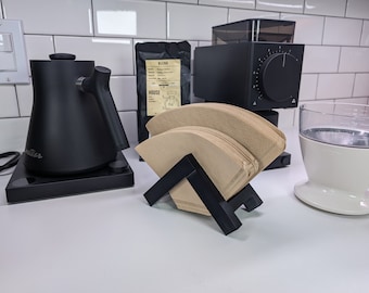 May include: A black coffee maker with a black kettle, a black grinder, and a white ceramic pour-over coffee dripper. The coffee maker is on a white countertop with white tile backsplash. There are brown coffee filters in a black holder.