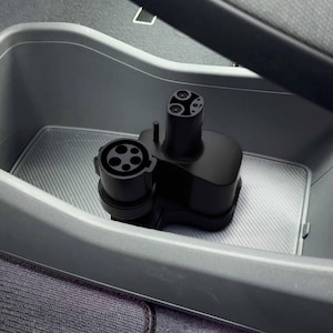 May include: A black electric vehicle charging adapter featuring a circular connector with five pin holes on one side and a smaller, elongated connector with two pin holes on the top, designed for connecting different types of charging stations.