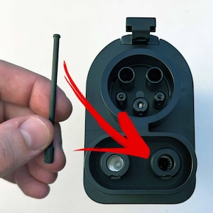 May include: A black electric vehicle charging port with multiple pin receptacles. A small, black, cylindrical tool is held nearby, possibly for maintenance or cleaning. A red arrow points to a specific port.