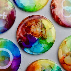 May include: Seven round, colourful, resin ornaments with white symbols. The symbols are a circle, a transgender symbol, a lesbian symbol, and a gay symbol. The ornaments are painted with shades of blue, green, yellow, orange, red, and purple.