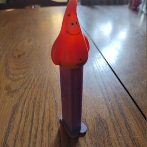 May include: A purple and red plastic toy with a cartoon character head. The character is red and has a smiling face. The toy is a dispenser for candy.