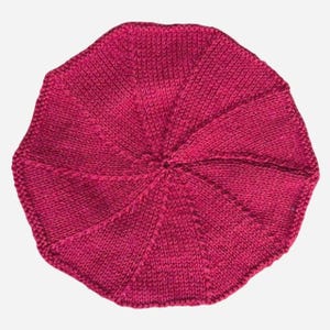 May include: A handmade, knitted beret in a vibrant shade of fuchsia. The beret features a circular design with radiating segments, creating a textured appearance. The hat is made of a soft, warm yarn, perfect for cooler weather.