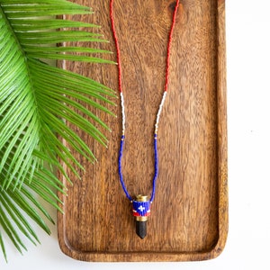 May include: A beaded necklace with a bullet casing pendant. The bullet casing is wrapped with blue, white and red beads, resembling a flag. The necklace is displayed on a wooden tray.