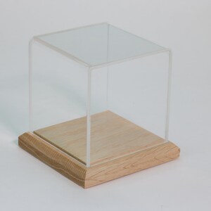 Puede incluir: Una vitrina de exhibici&oacute;n de cubo de acr&iacute;lico transparente con una base de madera. La base es de madera clara y tiene un acabado liso.