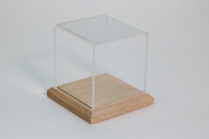 Puede incluir: Una vitrina de exhibici&oacute;n de cubo de acr&iacute;lico transparente con una base de madera. La base es de madera clara y tiene un acabado liso.