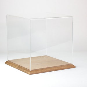 Puede incluir: Una vitrina de exhibición de acrílico transparente con una base de madera. La vitrina es de forma cúbica y tiene una base de madera clara.
