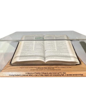 May include: A wooden box with a glass lid, displaying an open book. The box has a plaque with the inscription "In Memory of Fredrick Chikwendu also known as F.C. Kolo A loving husband, a beloved father, a man of deep respect and unwavering faith, a devoted servant of God."