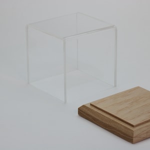 Puede incluir: Una vitrina de cubo de acr&iacute;lico transparente con una base de madera. La vitrina est&aacute; abierta y la base est&aacute; separada.
