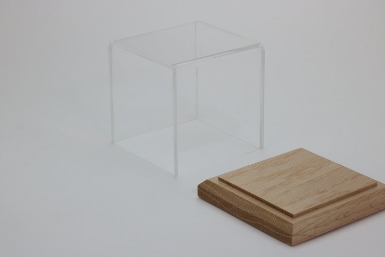 Puede incluir: Una vitrina de cubo de acr&iacute;lico transparente con una base de madera. La vitrina est&aacute; abierta y la base est&aacute; separada.