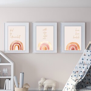 Puede incluir: Tres impresiones de arcoíris de acuarela enmarcadas con citas inspiradoras en letras doradas. Las impresiones están colgadas en una pared beige claro sobre una estantería blanca y una tienda de campaña tipi azul y blanca.