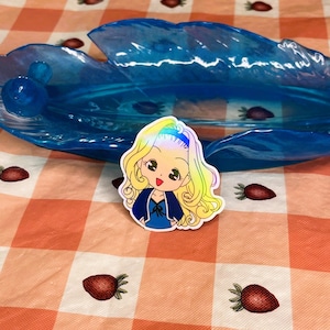 May include: A holographic sticker of a cartoon girl with blonde hair, wearing a blue dress and a white collar. The sticker is on a pink and white checkered background.