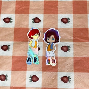 May include: Two holographic stickers of cartoon characters. The characters are both young people with short hair. One character has red hair and is wearing a yellow shirt and blue shorts. The other character has brown hair and is wearing an orange shirt and white shorts.