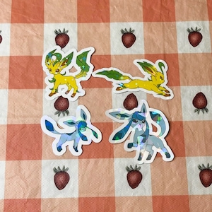 May include: Four holographic stickers featuring different forms of the Pokemon Eevee. The stickers are on a pink and white checkered background.