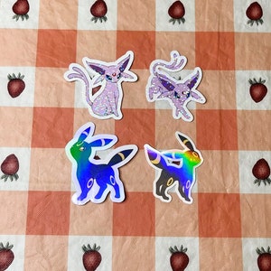 May include: Four holographic Pokemon stickers featuring Espeon and Umbreon. The stickers are on a pink and white checkered background.