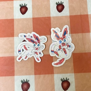 May include: Two sparkly holographic stickers of the Pokemon Sylveon, one in a standing pose and the other in a sitting pose. Both stickers are on a pink and white checkered background.