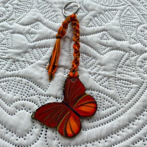 May include: A red and orange butterfly keychain with a braided orange and brown tassel. The butterfly is painted with a detailed design and has a silver key ring.