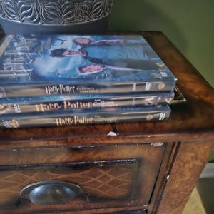 May include: Three Harry Potter DVD movie cases stacked on a wooden table. The top case features a blue and white cover with actors. The other two cases are black with the movie titles.