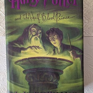 May include: A hardcover book titled "Harry Potter and the Half-Blood Prince" with a green and purple cover. The cover features illustrations of Dumbledore, Harry Potter, and a glowing bowl. The author's name, J.K. Rowling, is at the bottom.