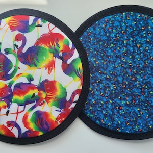 May include: Two round fabric items with black trim. One features a white background with colorful flamingos. The other has a blue background with a speckled pattern of various colors. These items could be coasters or trivets.