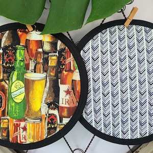 以下が含まれることがあります： 2つの丸い布製鍋敷き。1つは、さまざまな色のビール瓶とグラスのコラージュが特徴です。もう1つは、白の背景に黒とグレーのシェブロンの繰り返し模様があります。どちらも黒い縁取りがあります。