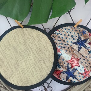May include: Two round trivets. One has a beige, textured pattern, and the other features a patriotic design with red, white, and blue stars on a wood-grain background. Both have a black border.