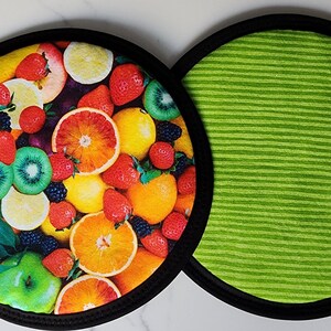 May include: Two round trivets. One features a vibrant fruit medley of strawberries, oranges, lemons, kiwi, and blackberries. The other has a green striped pattern. Both have a black border.