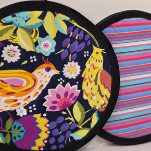 May include: Two round fabric pot holders with black trim. One features a dark blue background with yellow, orange, and pink birds and flowers. The other displays horizontal stripes in shades of pink, blue, purple, and white across the entire surface.