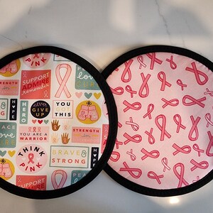 Op de afbeelding: Twee ronde stoffen items met zwarte rand. Eén heeft een patroon van roze linten op een roze achtergrond. De andere heeft een witte achtergrond met roze linten en teksten als "Think Pink" en "Brave & Strong".