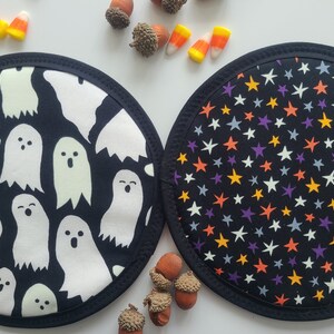 Könnte beinhalten: Zwei runde Halloween-Topflappen. Einer zeigt weiße Geister-Designs auf schwarzem Hintergrund, der andere bunte Sterne auf schwarzem Hintergrund. Beide haben einen schwarzen Rand. Süßigkeiten und Eicheln sind verstreut.