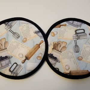 Puede incluir: Dos salvamanteles redondos de tela con borde negro. La tela azul claro tiene un estampado de utensilios de repostería vintage, recetas, huevos y bayas. Se ven un rodillo, una batidora y cucharas de madera.