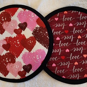 May include: Two round fabric pot holders. One features a pattern of red, pink, and burgundy hearts on a white background. The other has a burgundy background with the word "love" and small hearts. Both have black trim.