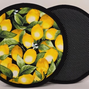 May include: Two circular pot holders with black trim. One features a vibrant pattern of yellow lemons and green leaves on a black background. The other shows a black underside with a white dotted, non-slip texture for grip and heat protection.