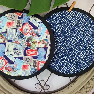 May include: Two round pot holders. One features a collage of vintage U.S. postage stamps with patriotic themes, including the Statue of Liberty and portraits of historical figures. The other has a blue and white grid pattern. Both have black trim.