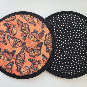 May include: Two round fabric trivets. One features a pattern of orange and black monarch butterflies on an orange background. The other has a black background with white polka dots. Both have a black border.