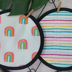 May include: Two round pot holders with black trim. One has a white background with repeating rainbow graphics. The other has a white background with horizontal stripes in shades of pink, blue, and yellow.