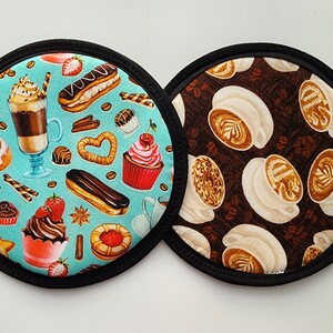 May include: Two round fabric pot holders. One has a teal background with coffee and pastry illustrations, including a latte, cupcake, and eclair. The other has a brown background with coffee art illustrations. Both have black trim.