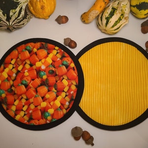 Puede incluir: Dos manteles individuales redondos con ribete negro. Uno presenta un patrón de caramelos de maíz y calabazas, el otro tiene un diseño de rayas verticales amarillas. Calabazas y bellotas están esparcidas alrededor de los manteles individuales.