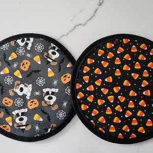 May include: Two round Halloween-themed pot holders. One features a gray background with cartoon dogs, pumpkins, bats, candy corn, and spiderwebs. The other has a black background with candy corn.