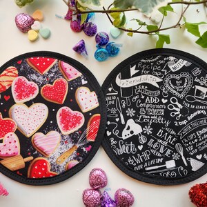 May include: Two round black fabric coasters. One features heart-shaped cookies with red and white icing. The other has a recipe for friendship with text and illustrations in white. Valentine's Day theme.