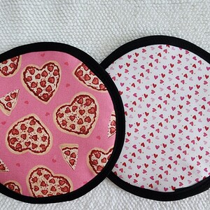 May include: Two round fabric pot holders with black trim. One features a pink background with heart-shaped pizzas and pizza slices. The other has a white background with red and pink hearts.