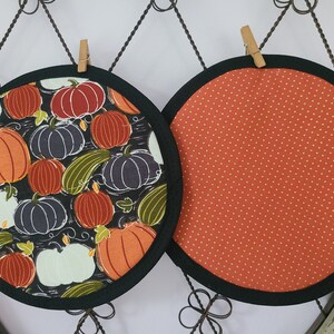 May include: Two circular quilted pot holders. One has a black background with orange, white, and navy pumpkins and green gourds. The other has an orange background with white polka dots. Both have black trim.